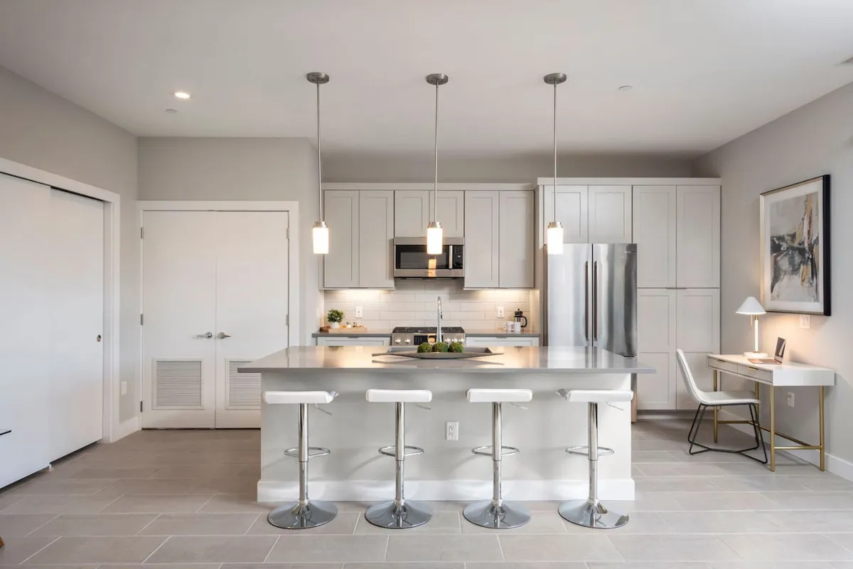A bright and contemporary kitchen with a spacious island surrounded by bar stools, stainless steel appliances, and vibrant artwork on the wall. The space is open and inviting, ideal for gathering with friends or preparing meals before heading out. Natural light enhances the welcoming mood, hinting at the social possibilities connected to the central location.