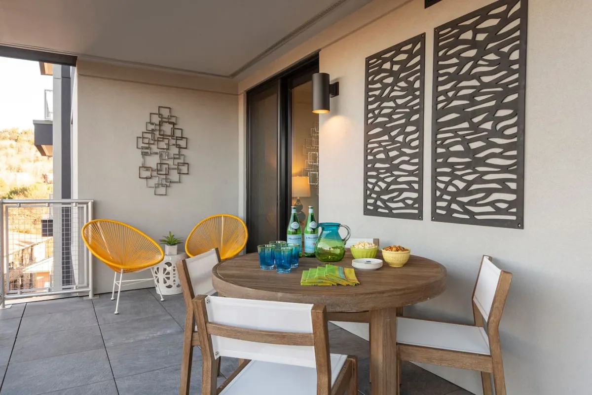 An exterior scene showing a stylish round wooden table with yellow Acapulco chairs and decorative wall art, set for outdoor dining with glassware and food items. The space has an open, welcoming ambiance, indicating a transition from private parking or entry to the comfortable living space.