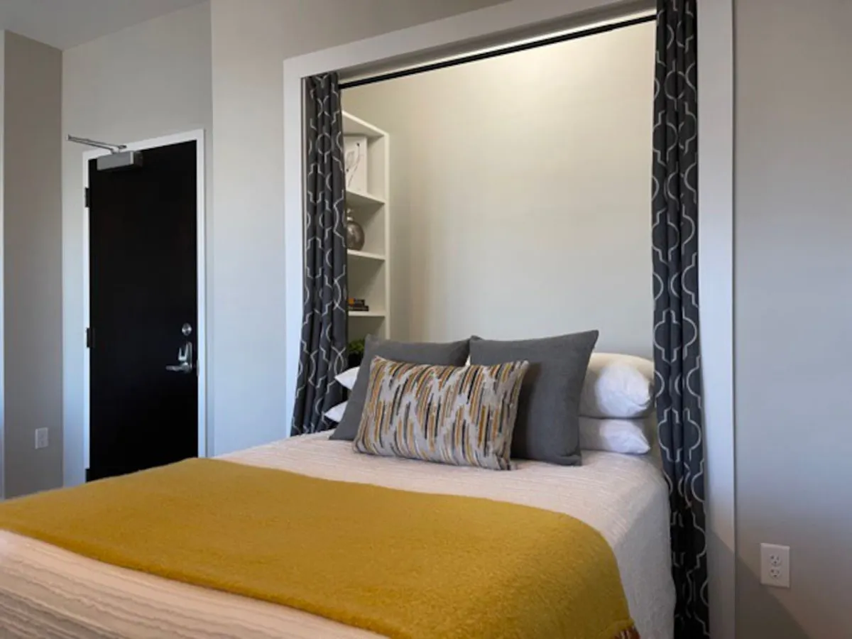 A cozy and peaceful bedroom setting with a bed adorned in a yellow blanket and decorative pillows, complemented by curtains framing the sleeping area. The mood is calm and restful, ideal for reading or unwinding.