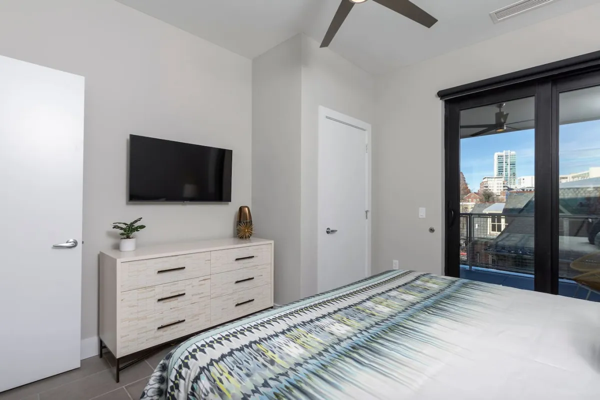 This bedroom scene includes a bed with a patterned comforter, television, and dresser with a decorative plant. The angle and staging suggest the bedroom opens onto the balcony, reinforcing its accessibility and the opportunity to enjoy scenic views right from bed.