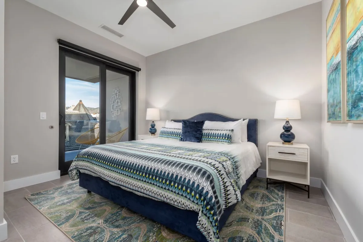 A serene and comfortable main bedroom featuring a bed with patterned bedding, abstract artwork above the headboard, nightstands with lamps, and large windows leading directly to a balcony. Sunlight streams through the windows, creating a soft, inviting atmosphere perfect for relaxation, while the balcony access highlights flexibility and indoor-outdoor living.