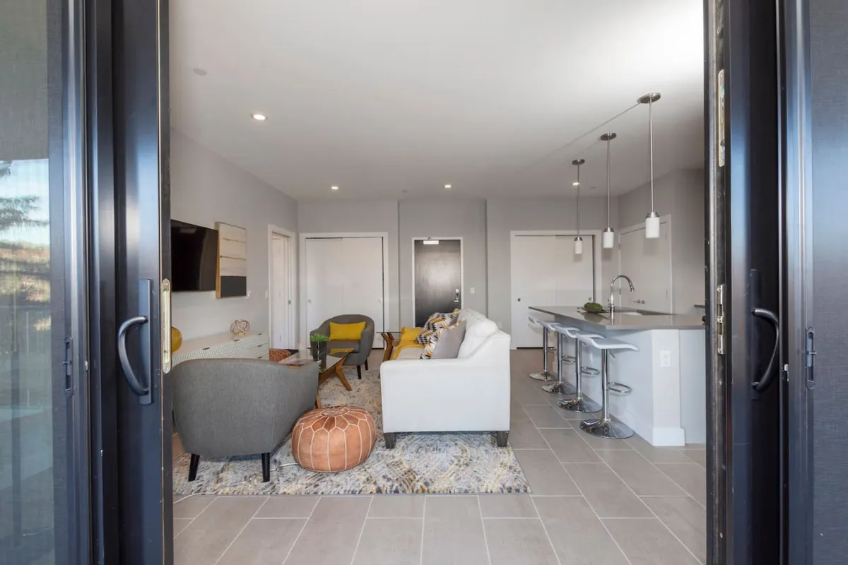 A contemporary living and dining area with a comfortable sofa, bar stools along the kitchen counter, a stylish rug, curated wall decor, and abundant natural light.