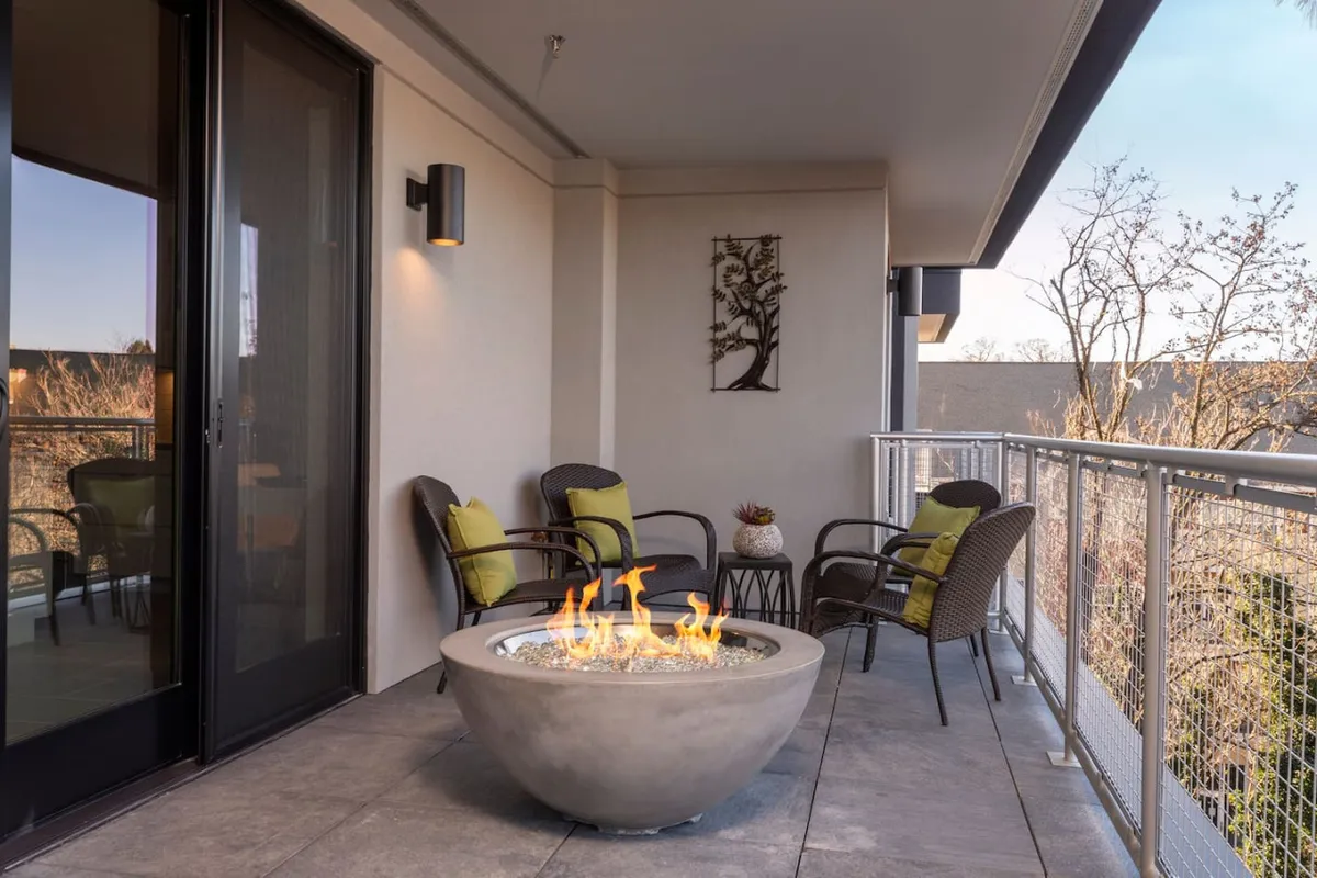 An inviting balcony setup featuring a fire pit surrounded by stylish chairs and accent decor. The backdrop includes a city skyline visible at dusk, creating a warm, atmospheric setting for socializing or quiet reflection.
