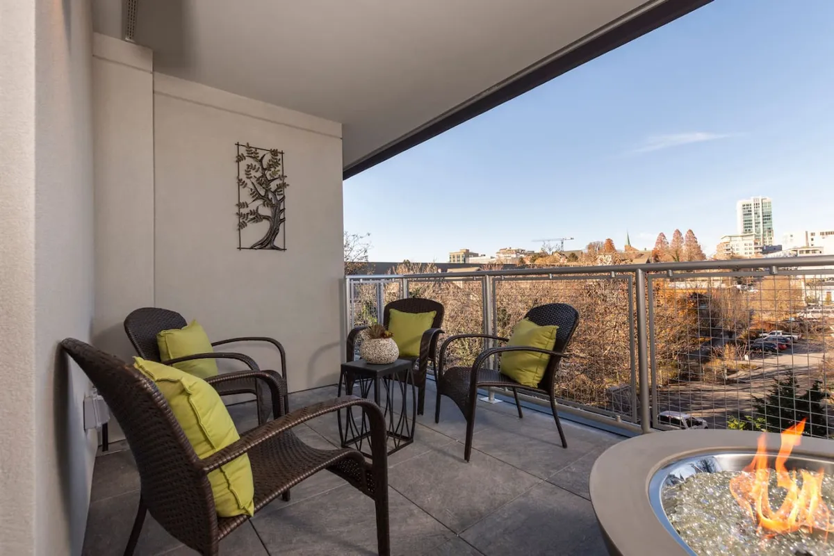This exterior image showcases a spacious balcony furnished with comfortable wicker chairs, a modern fire pit, decorative wall art, and a sweeping city view beyond. The scene is set for both daytime relaxation and cozy evenings, with the city skyline and mountain air visible from the outdoor lounge area.