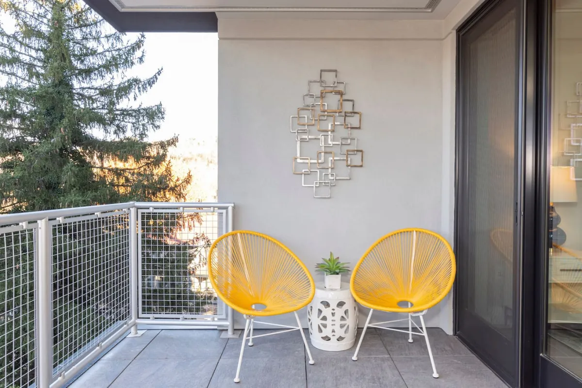 A cheerful, cozy balcony scene with vibrant yellow chairs, decorative wall art, and lush potted plants. The setup suggests mornings spent sipping coffee while soaking in natural light and city energy.