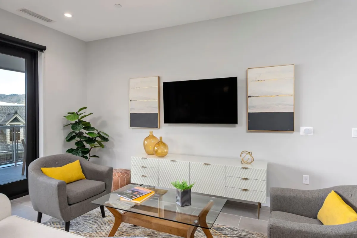 The image features a contemporary living area with a television, artwork, plant, and a coffee table. Large windows in the background suggest a connection to an outdoor balcony, hinting at sunset views and the indoor-outdoor lifestyle described in the section.
