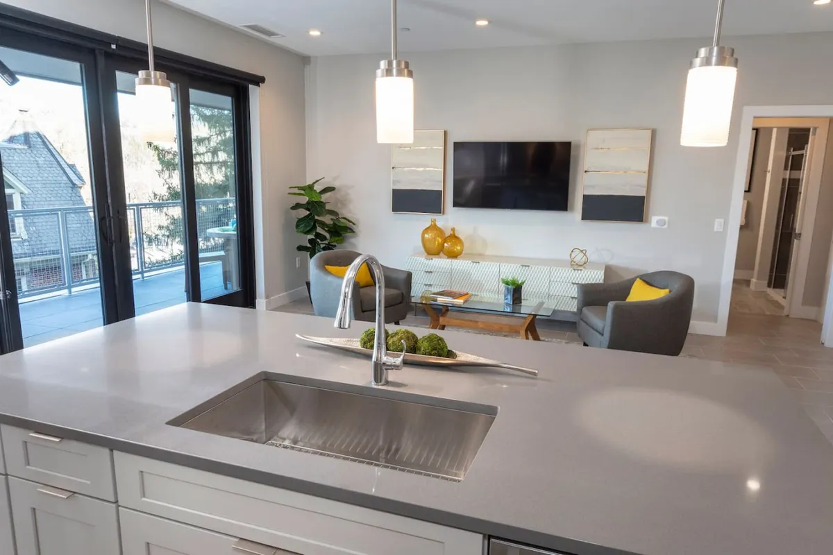 A contemporary open-plan living area featuring a kitchen island with seating, a lounging space, television, and decorative items. The arrangement invites both relaxation and casual entertaining, with a glimpse of the balcony area hinting at indoor-outdoor living.