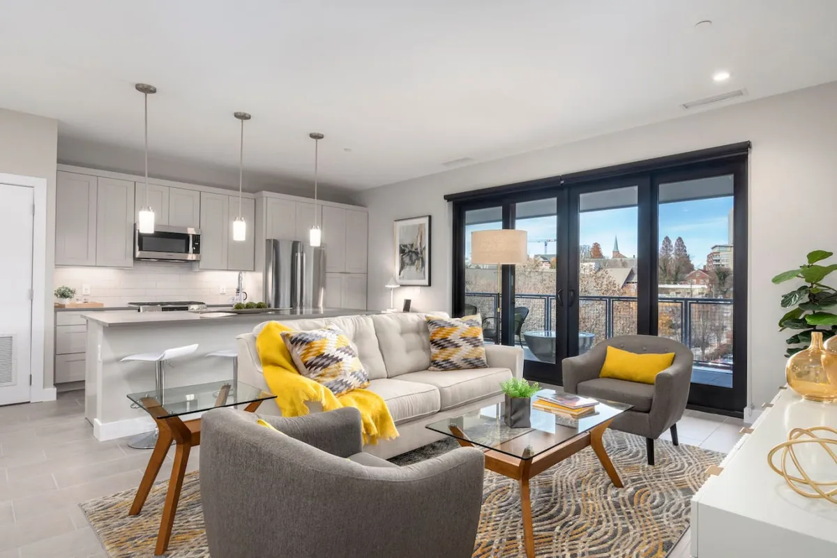 An open-concept living and kitchen space featuring a plush sofa with a yellow throw, a glass coffee table, a modern kitchen in the background, and large windows that invite natural light and offer city views.